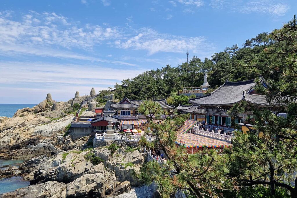 A temple complex near cliffs and the ocean on the left side and build into more of a forest on the right side.