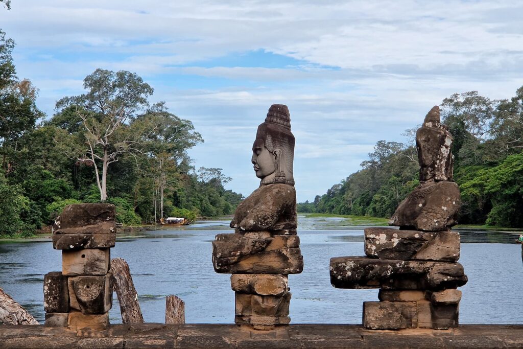 Old Statues lining on the bridge to the Tonle Om Gate surrounded by water in Angkor Wat.